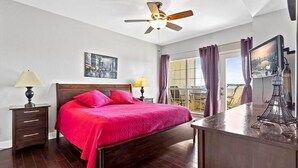 Een slaapkamer met een bed, nachtkastje, bureau, plafondventilator, raam met gordijnen en uitzicht op de stad.