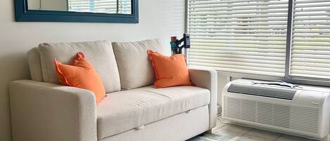 A living room with a beige sofa, two orange pillows, a large mirror, and a window with blinds.