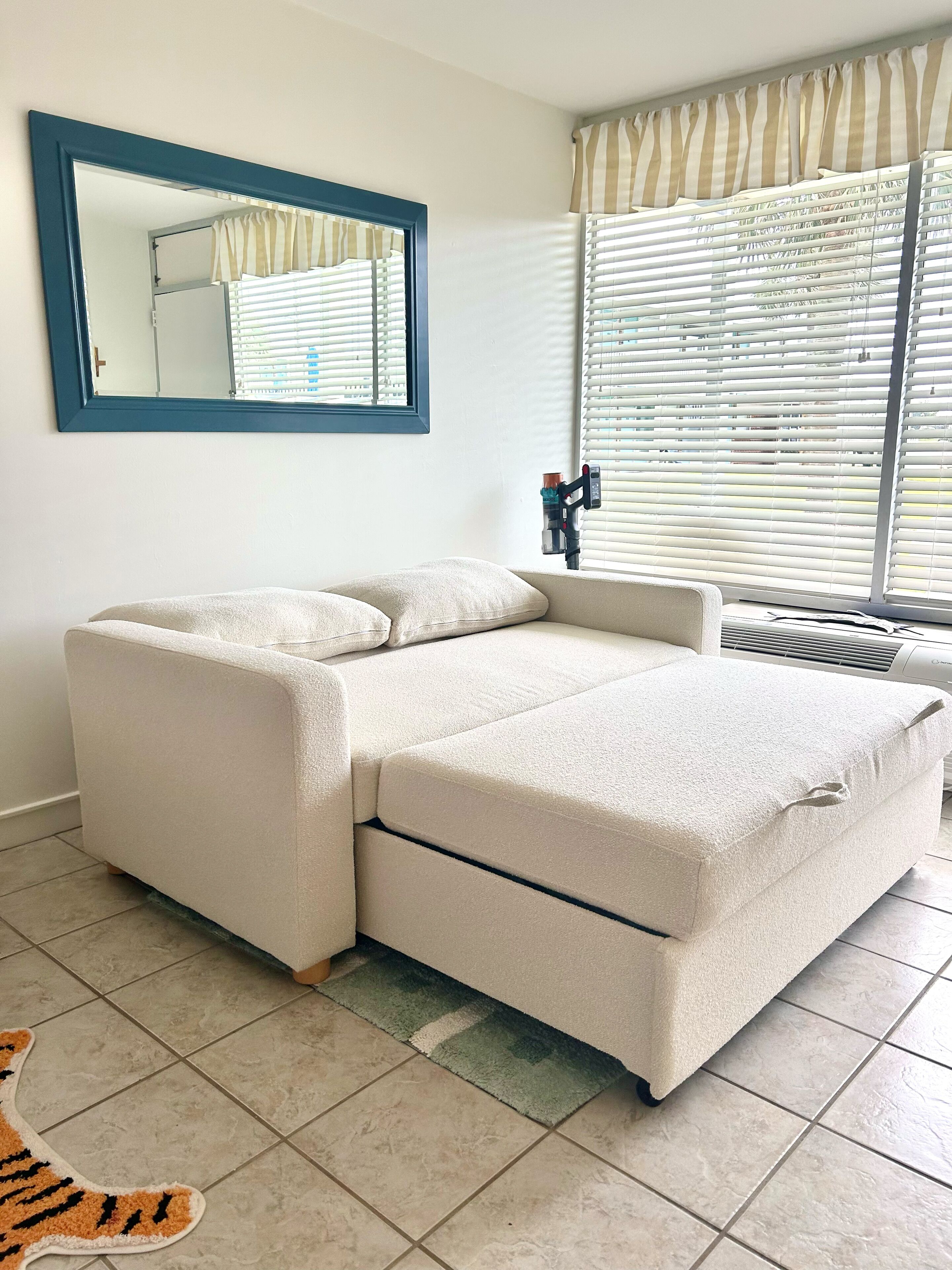 A modern living room with a beige sectional sofa, a large mirror, and a window with blinds.