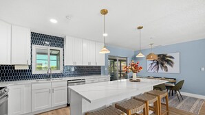 A modern kitchen with white cabinets, a large island with bar stools, pendant lighting, and a view of the outdoors. - One Last Summer Family Escape Pool• Arcade• Games (Bradenton)