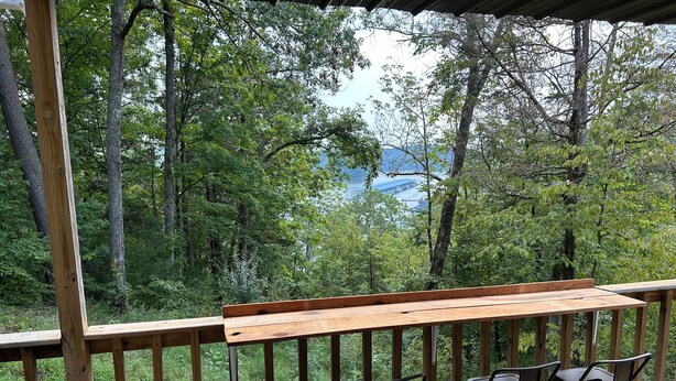 A wooden deck with a table and chairs, overlooking a forested area and a body of water. - Charming A-frame with Lake Cumberland Views (Jamestown)
