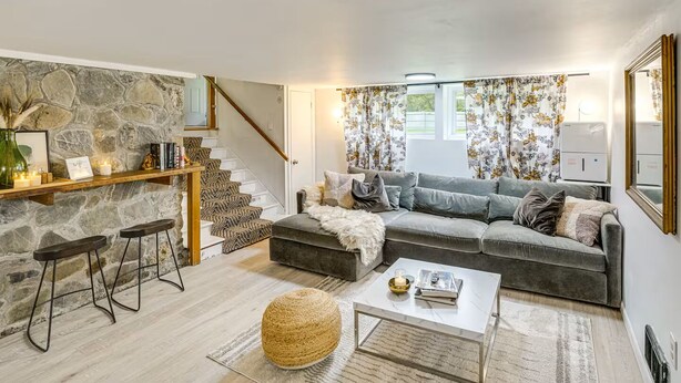A cozy living room with a stone wall, a grey sofa, a coffee table, and a staircase. - Private Cottage Close to Shopping, Skiing, Swimming, Hiking, Biking and Dining. (Austerlitz)