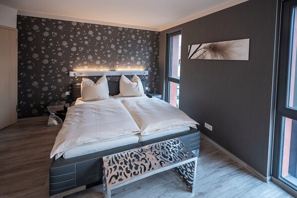 A modern bedroom with a large bed, a headboard with integrated lighting, a bench with a unique design, and a large window offering a view.