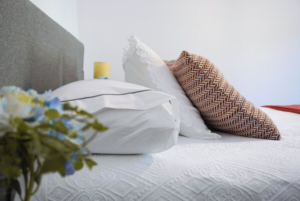 Una cama bien tendida con ropa de cama blanca, un cabecero gris y un cojín decorativo con diseño de espiga.