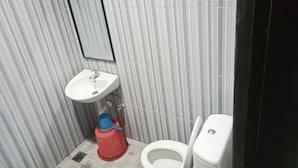 A small bathroom with a white sink, a mirror, a toilet, and a red cleaning bucket. - Hotel Taman Rai  (Tabanan)