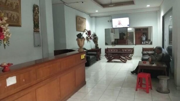 A reception area with a wooden counter, a sofa, a red stool, and a television mounted on the wall. - Hotel Taman Rai  (Tabanan)