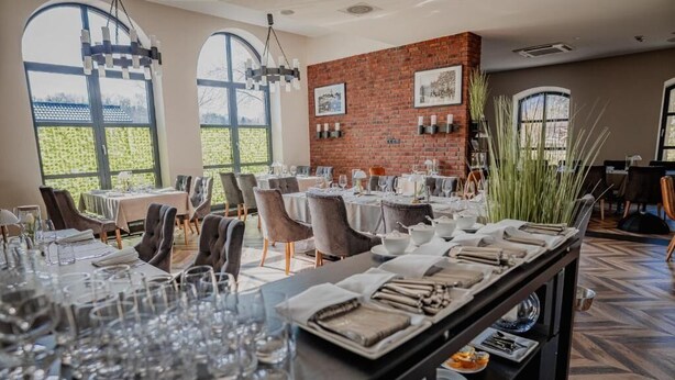 A restaurant with a long dining table set for a meal, large windows, and a brick wall. - Hotel Malbork Centrum Konferencyjne (Malbork)