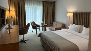 A hotel room with a bed, two armchairs, a desk with a lamp, a small table with a lamp, and a large window with curtains. - Hotel Malbork Centrum Konferencyjne (Malbork)