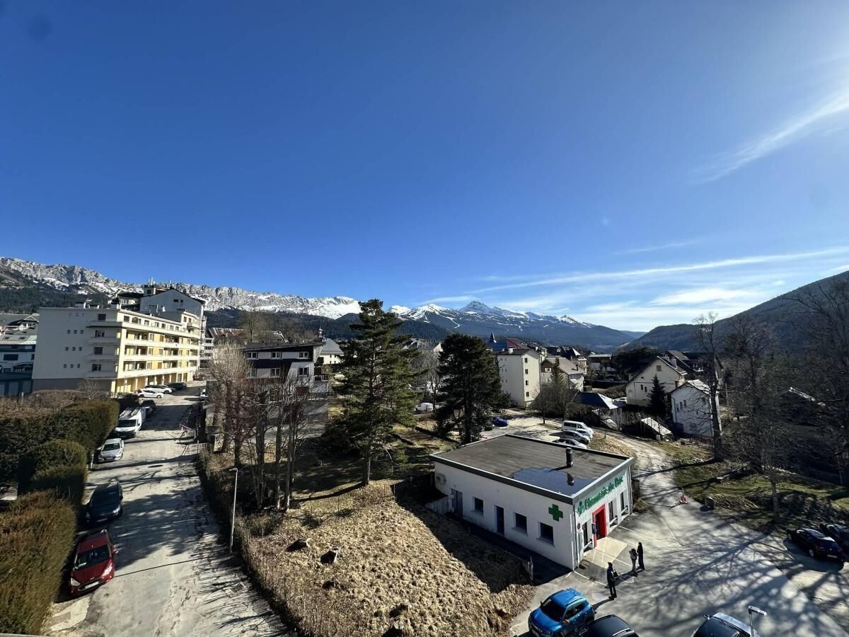 A building with a green cross sign, indicating a medical facility, surrounded by parked cars and a mountainous backdrop.