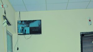 A reception desk with a television mounted above it, a door to the right, and a wall with some papers and a poster. - Hotel Narayan Palace (Danta Ramgarh)