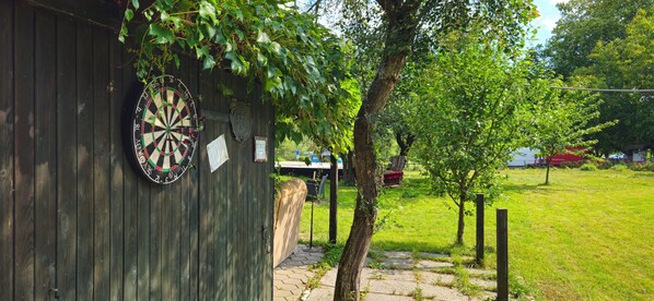 A dartboard mounted on a wooden wall, a pathway leading to a red building, and a grassy area with trees. - Maison du Danube - Nature, Culture, Relaxation & Sport (Aggsbach Markt)