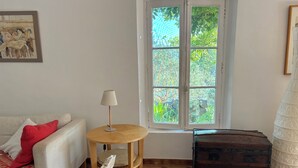 Une pièce avec un canapé blanc, une table en bois sur laquelle se trouve une lampe, une fenêtre donnant sur un paysage verdoyant et une vieille commode en bois foncé.