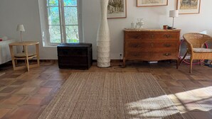 Une pièce avec une grande fenêtre, une malle en bois, un vase blanc, une chaise en osier et une commode à tiroirs.