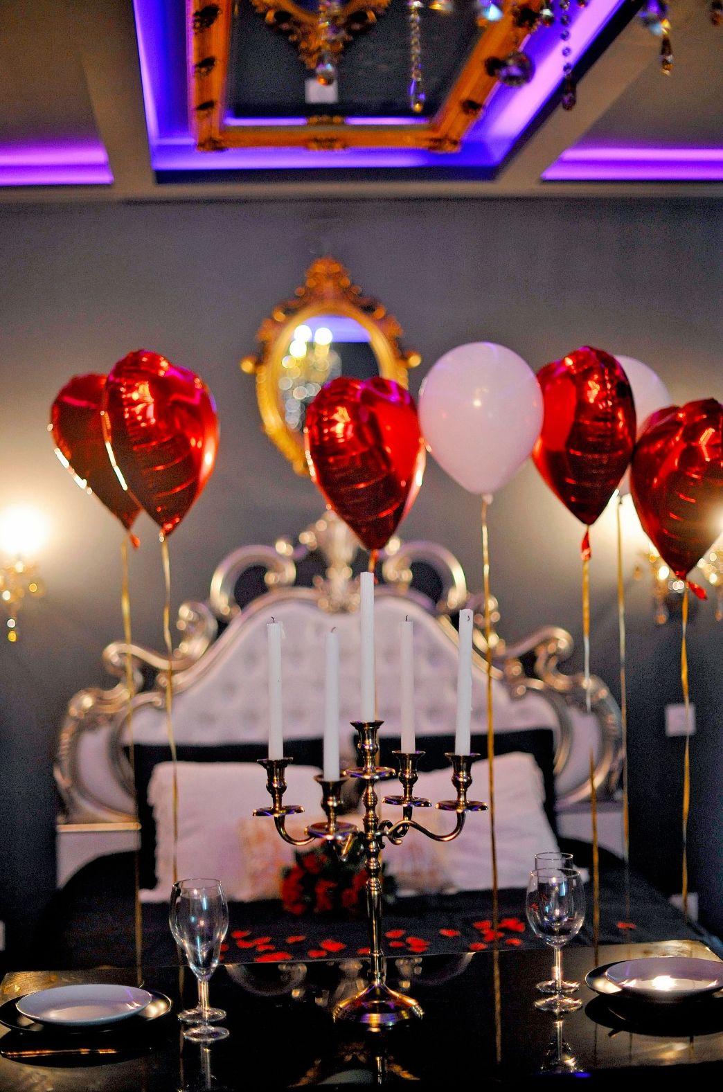 Una stanza con un letto, un candelabro, palloncini a forma di cuore e uno specchio appeso al muro.