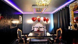 Une chambre luxueuse, ornée d’un lustre, avec un lit doté d’une tête de lit travaillée, une table sur laquelle est posé un vase de fleurs et des ballons en forme de cœur.
