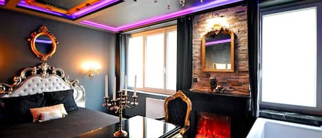 A luxurious bedroom with a large bed, ornate mirror, chandelier, and a fireplace.