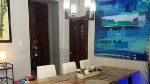 Un coin repas avec une table en bois, des chaises blanches et un banc. Un lustre est suspendu au-dessus de la table et un grand tableau abstrait orne le mur.