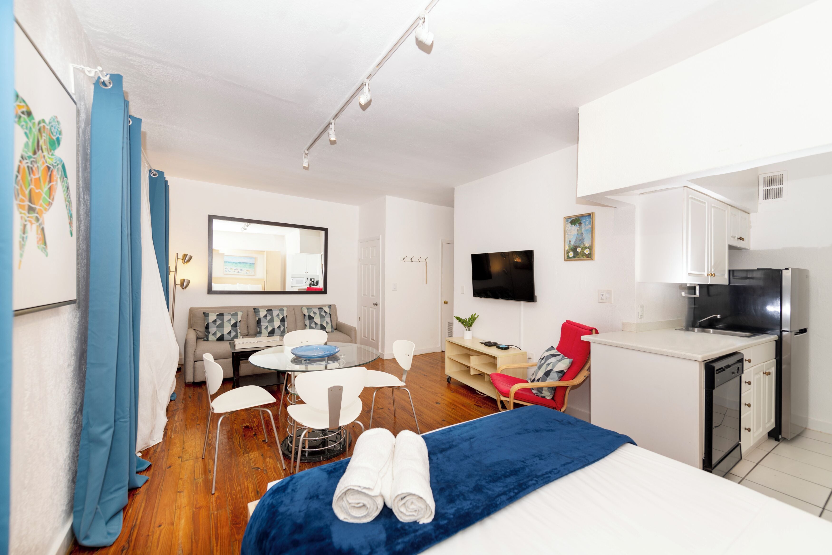 A modern hotel room with a bed, a small kitchenette, a dining area with a table and chairs, a sofa, and a TV mounted on the wall.