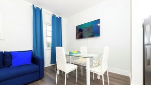 A modern living room with a blue sofa, white chairs, a glass dining table, and a kitchen area with a refrigerator. - Collins Beach Apartments (Miami Beach)