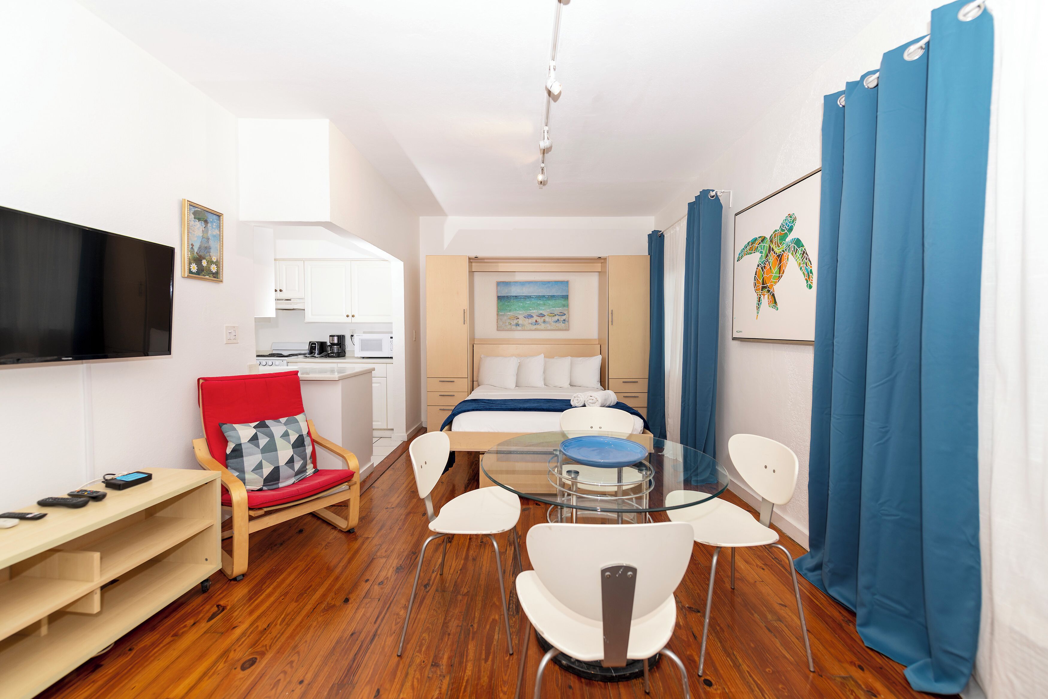 A compact hotel room with a bed, a small kitchenette, a dining area with a glass table, and a TV mounted on the wall.