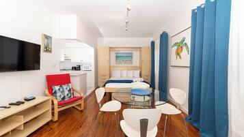 A compact hotel room with a bed, a small kitchenette, a dining area with a glass table, and a TV mounted on the wall.