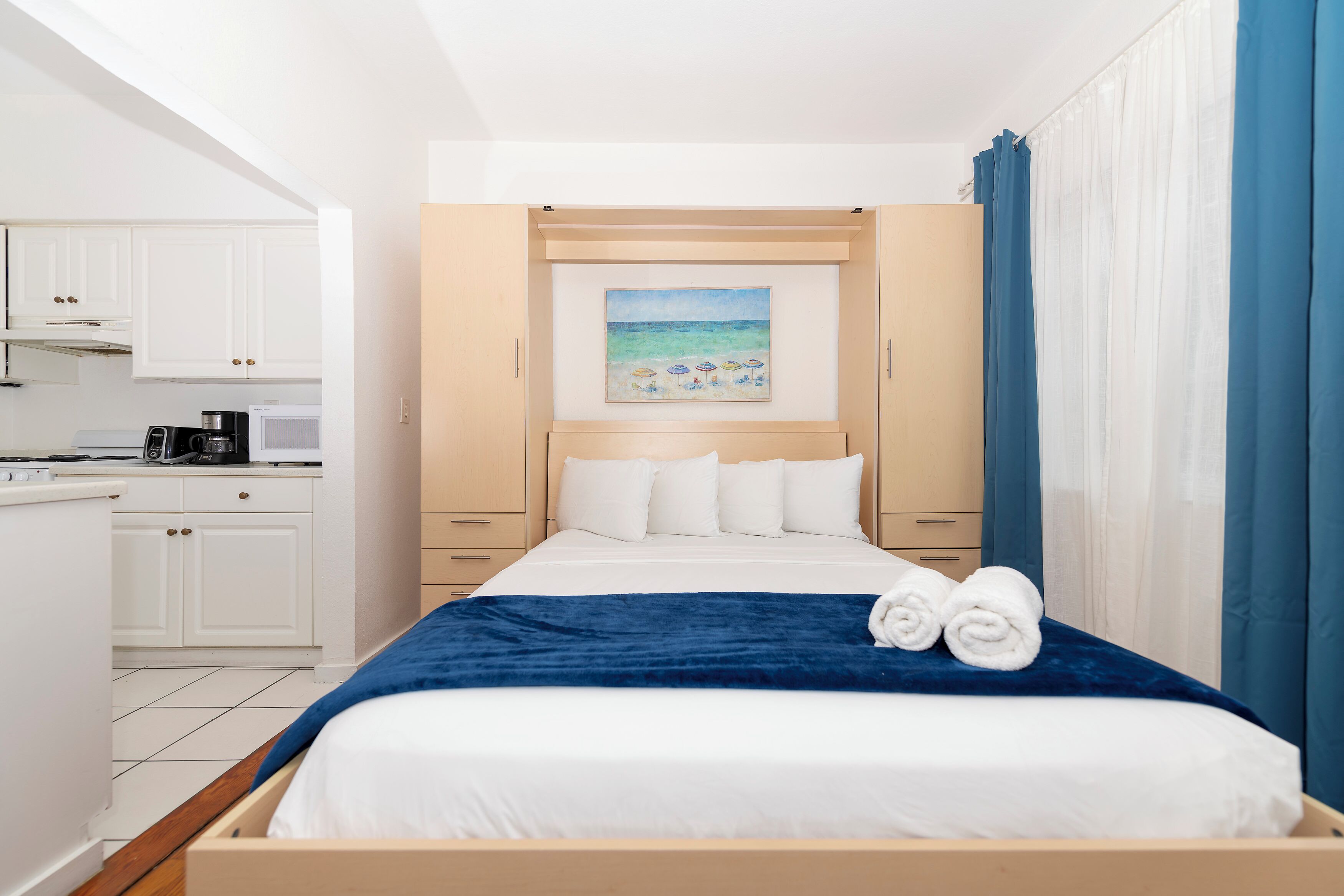 Ein Hotelzimmer mit einem Bett, einem Strandbild, einer Küchenzeile und einem Badezimmer mit blauen Vorhängen.