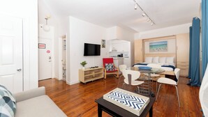 A modern living room with a sofa, a coffee table, and a dining area with chairs and a small table. There is a kitchen area with cabinets and a television. - Collins Beach Apartments (Miami Beach)