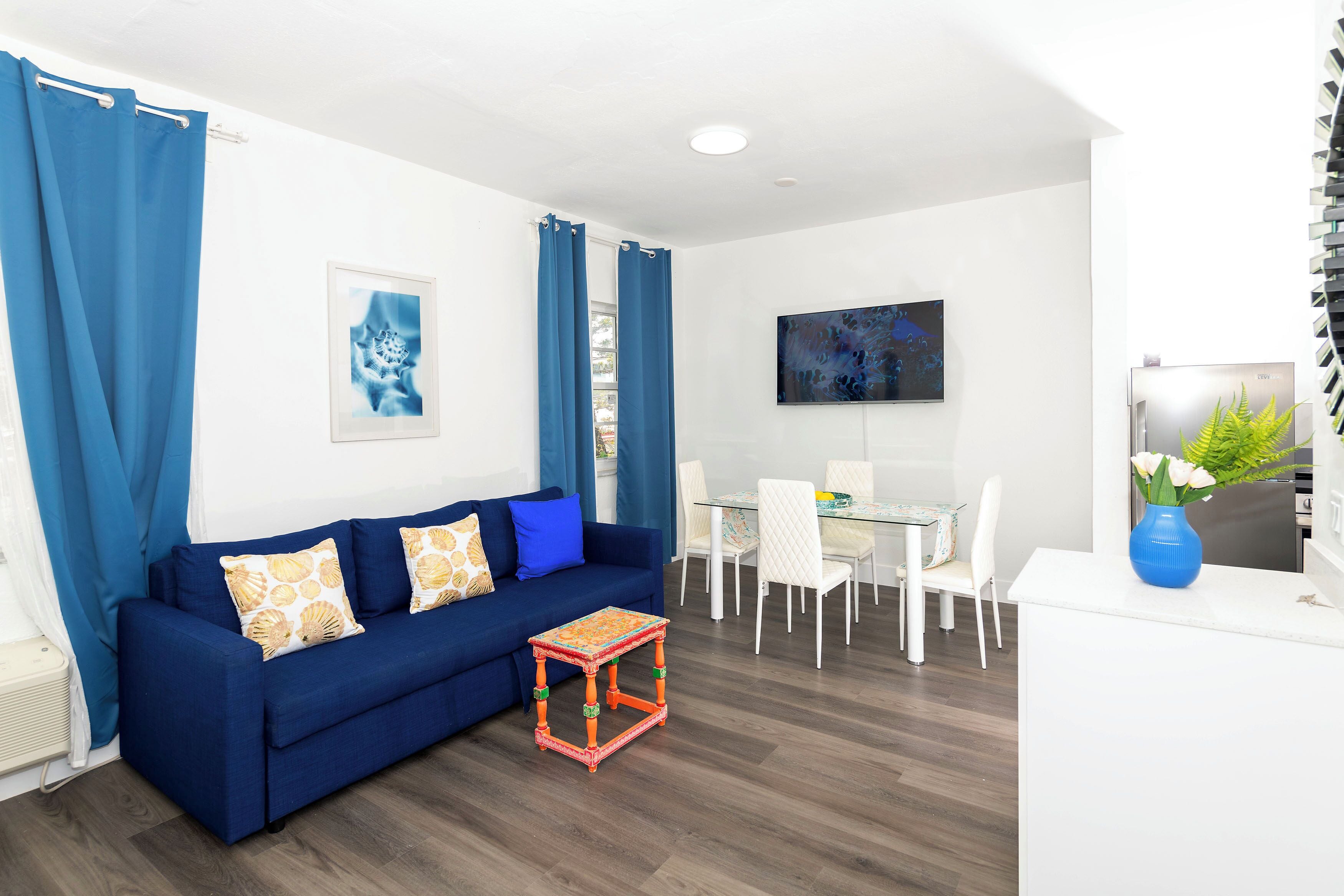 Una sala de estar moderna con un sofá azul, una mesita, un comedor con sillas y un televisor en la pared.
