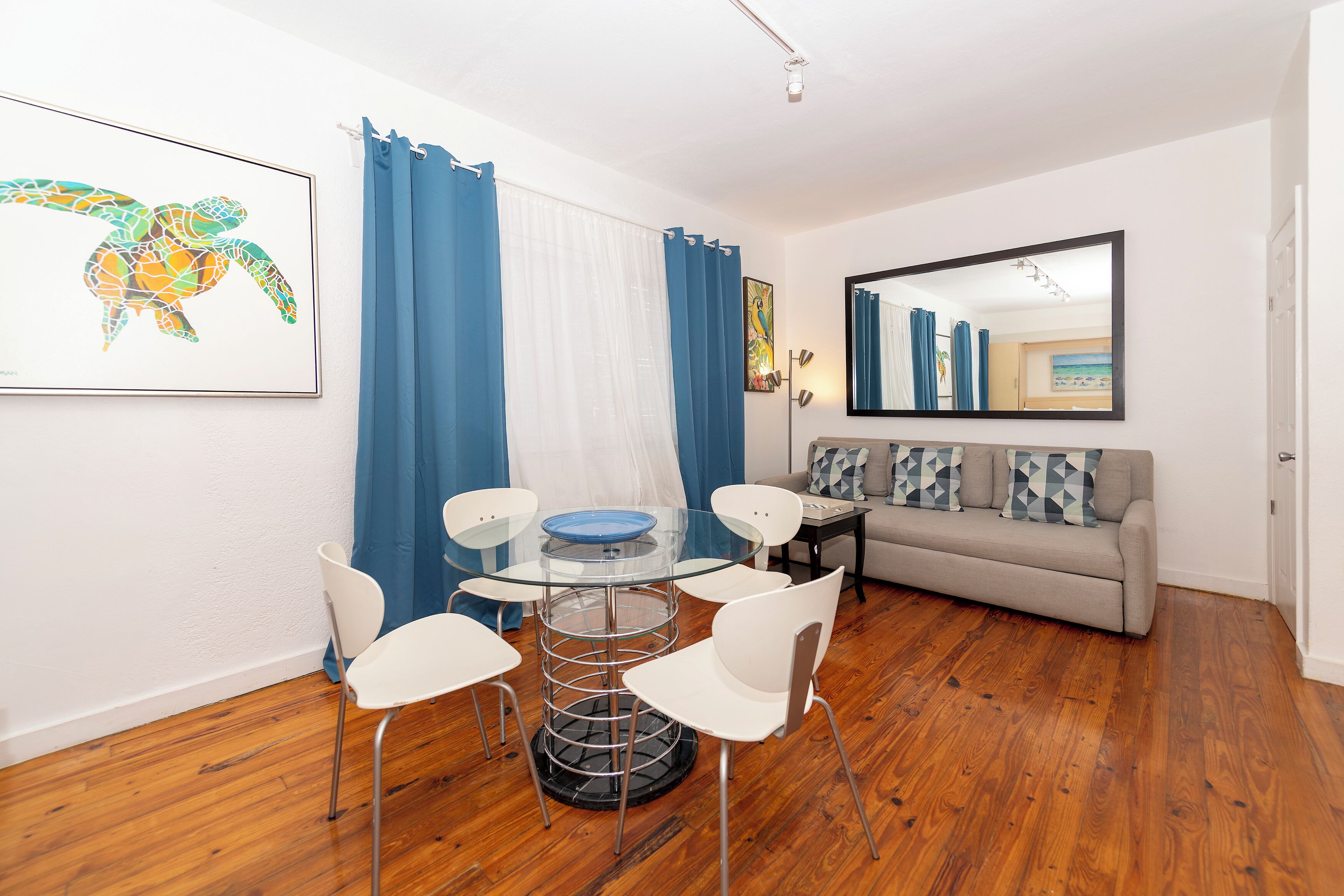 A modern living room with a glass table, white chairs, a sofa, and a large mirror.