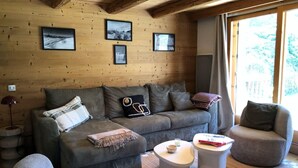 Een gezellig interieur van een chalet met een houten plafond, een bank, twee voetenbankjes en ingelijste foto’s aan de muur.