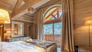 A cozy bedroom with a large bed, wooden ceiling beams, a window with curtains, and a view of the forest. - Chalet Altaïr with sauna, hot tub and hamam (Les Allues)