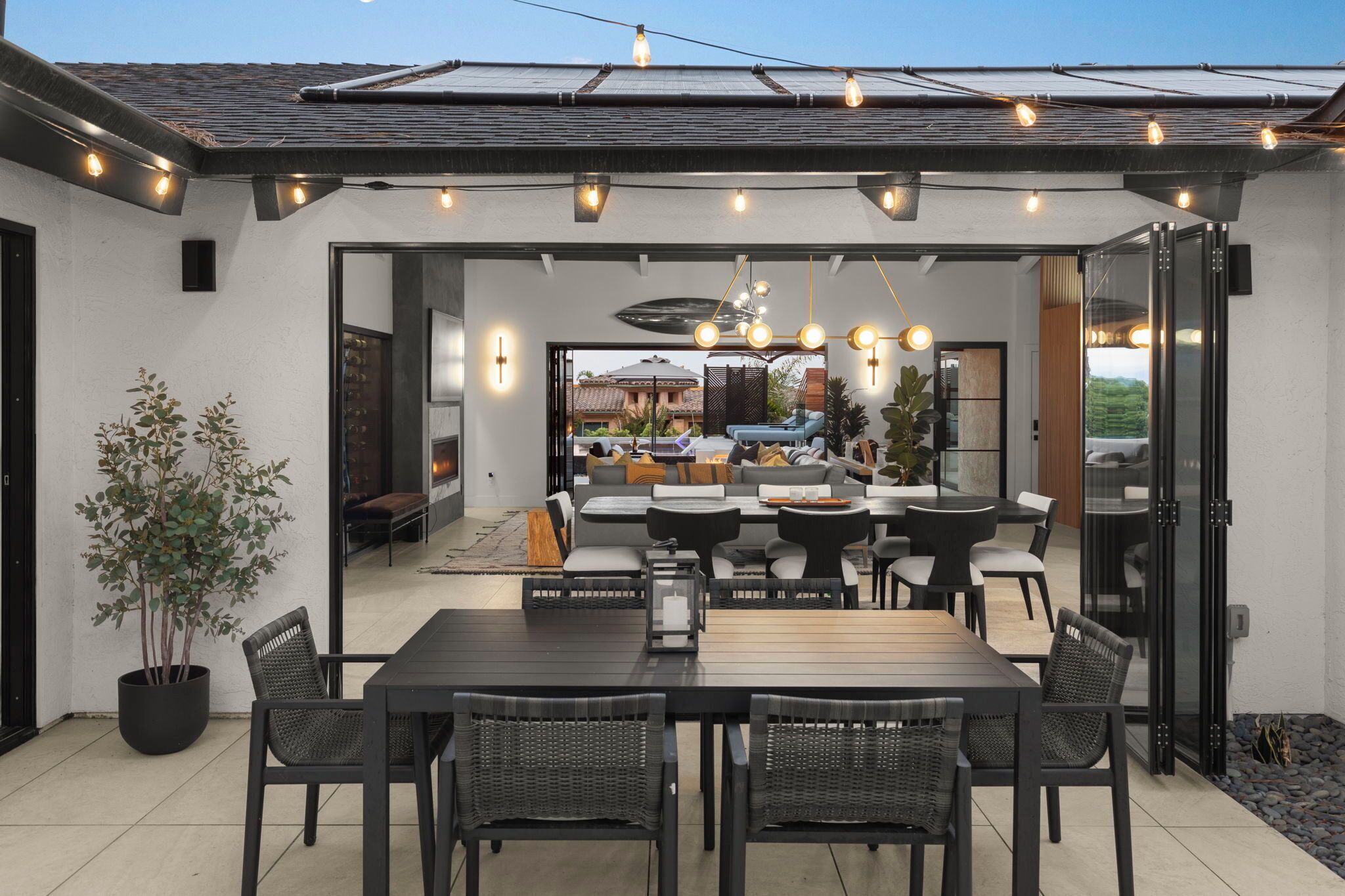 An outdoor dining area with a large table and chairs, a potted plant, and string lights overhead.