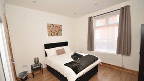 Un dormitorio con cama, mesita de noche, radiador, ventana con cortinas y un cuadro colgado en la pared.