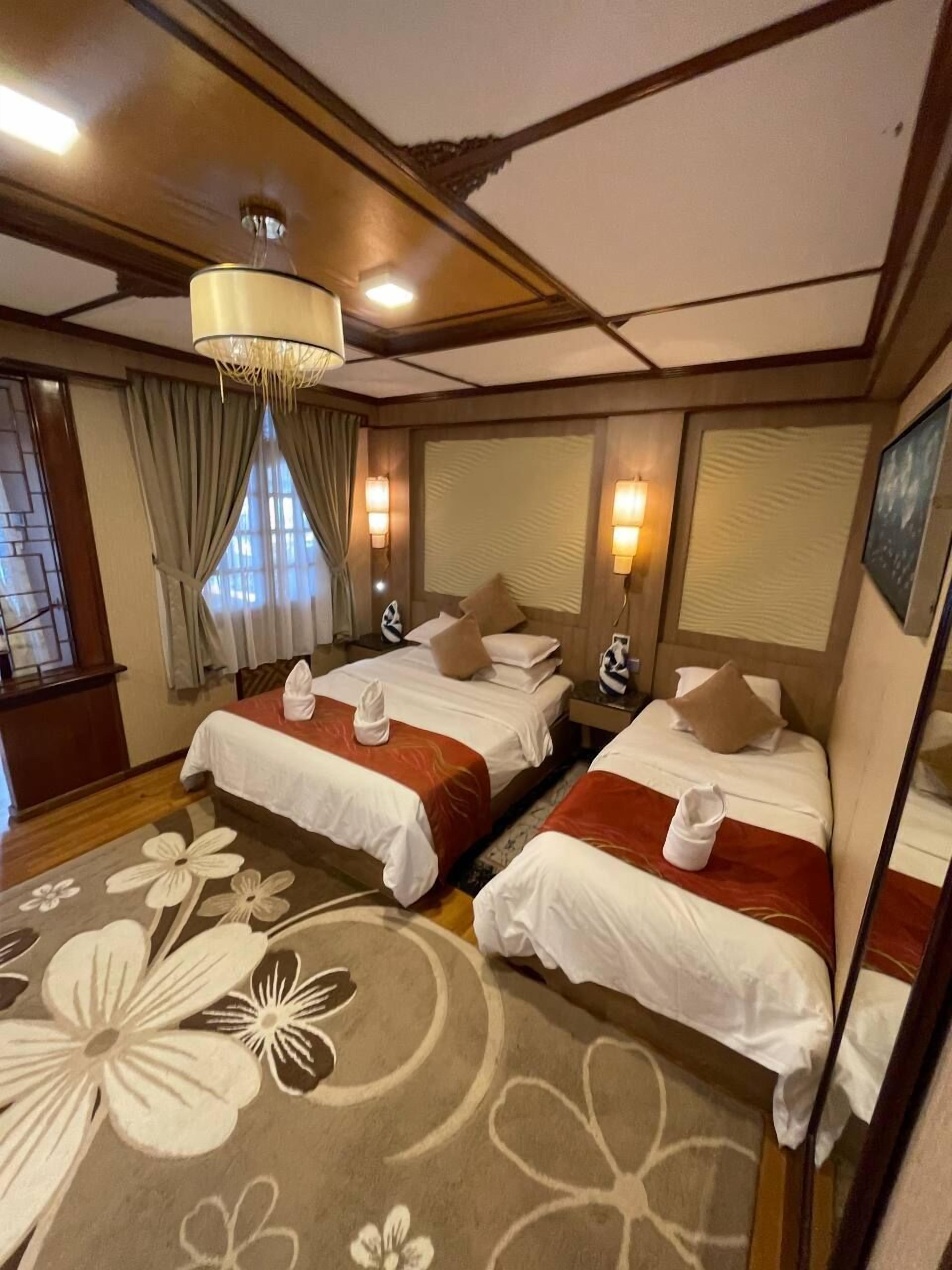 A hotel room with two beds, a chandelier, a window with curtains, and a floral carpet.