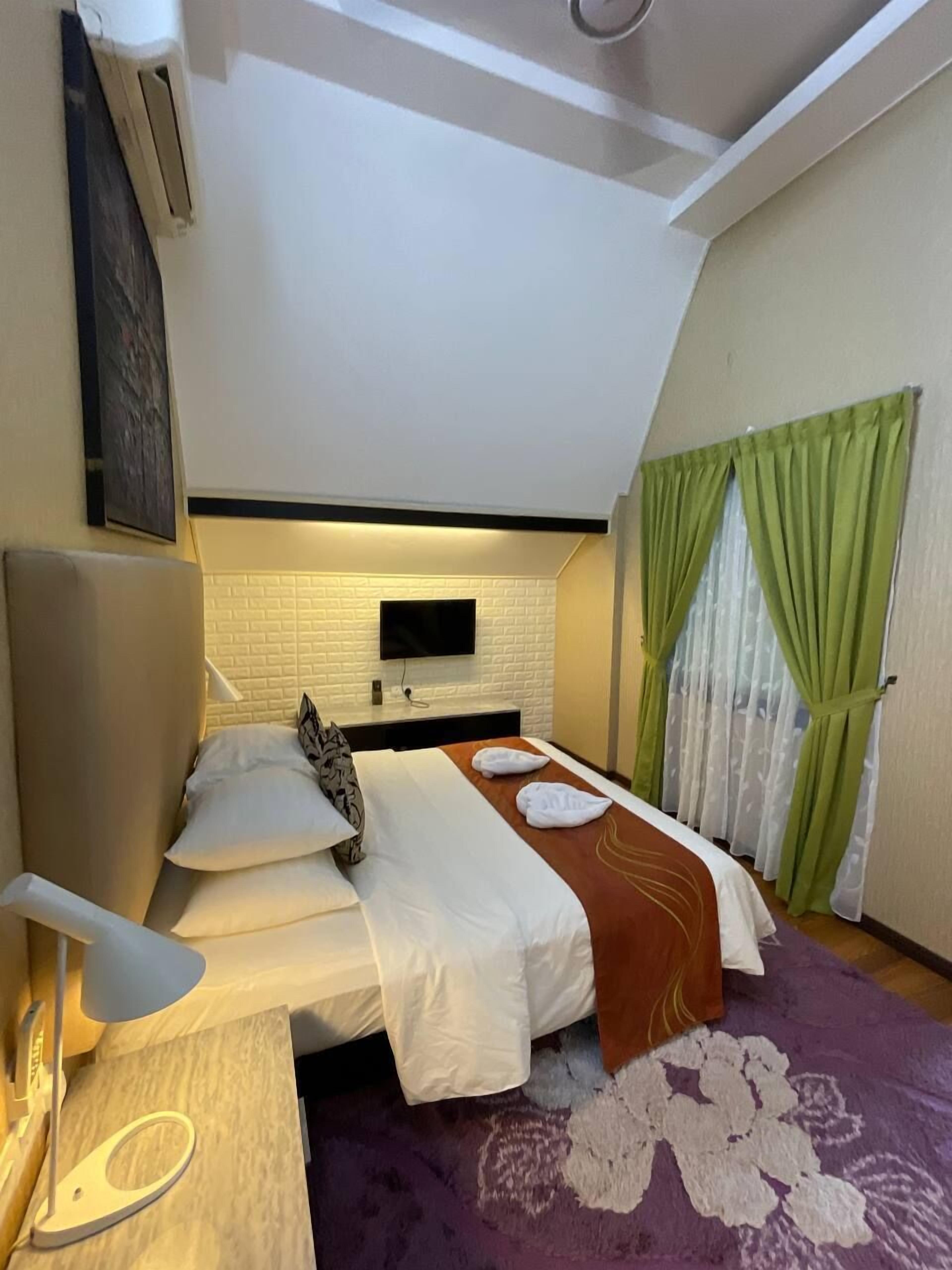A hotel room with a bed, a TV mounted on the wall, a desk with a lamp, and a window with green curtains.