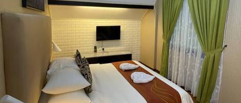 Une chambre d’hôtel avec un lit, un téléviseur fixé au mur, un bureau avec une lampe et une fenêtre avec des rideaux verts.