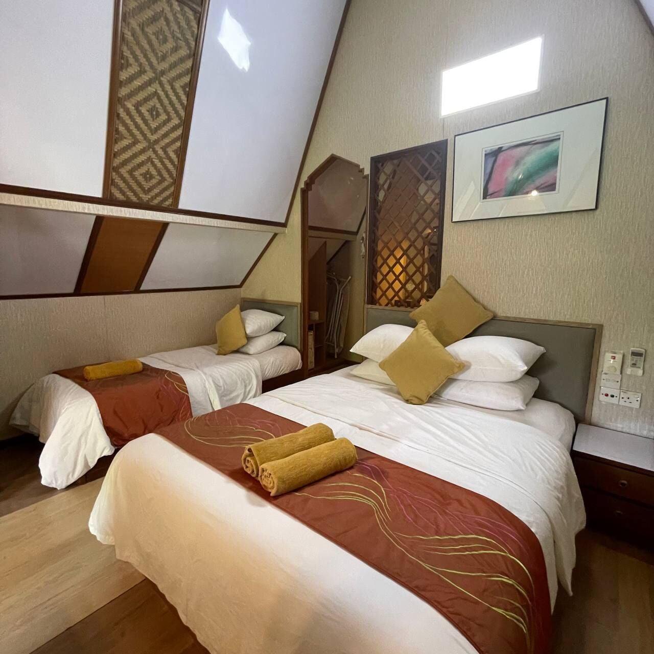 A room with two beds, each with a brown and white bedspread and pillows. There are two wall-mounted light fixtures and a framed picture on the wall.