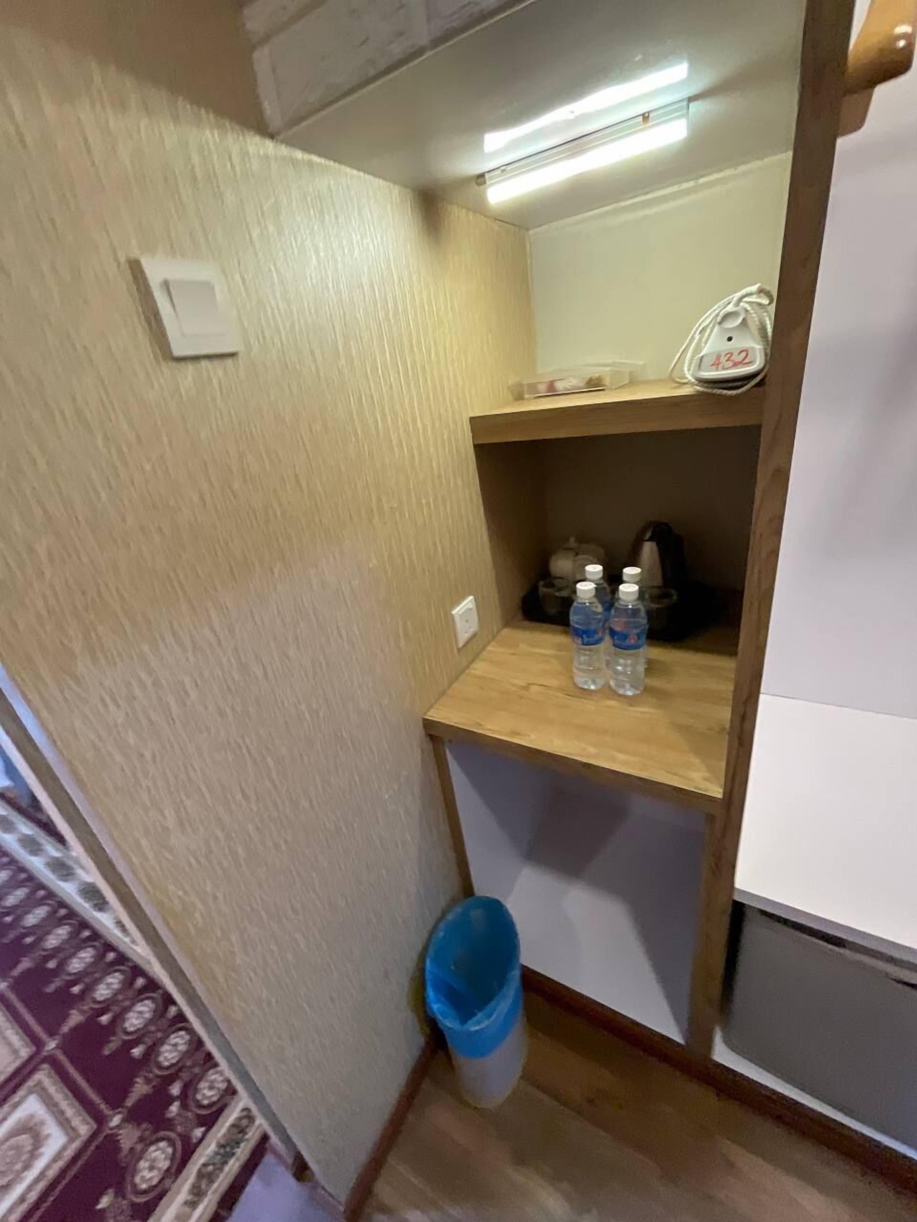 A small bathroom with a wooden shelf, a trash bin, and a white wall.