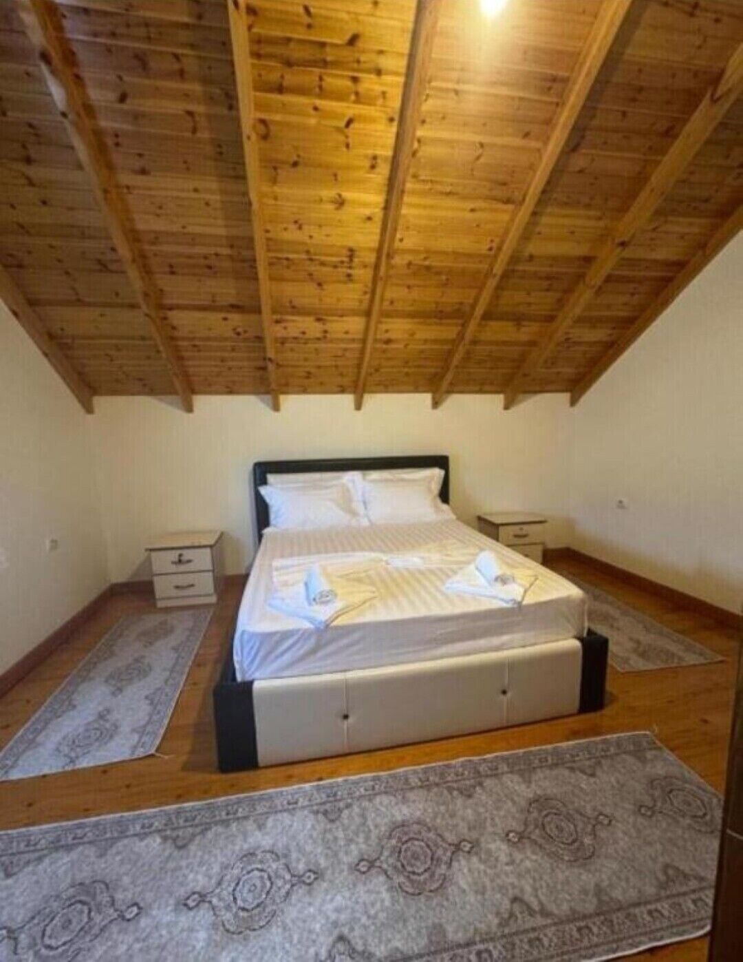 A bedroom with a wooden ceiling, a bed with a white mattress and pillows, two bedside tables, and a grey patterned rug.