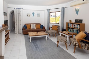 A living room with a brown sofa, patterned chairs, a wooden coffee table, a bookshelf, and a window with curtains. - Casa Lottie (Carvoeiro)