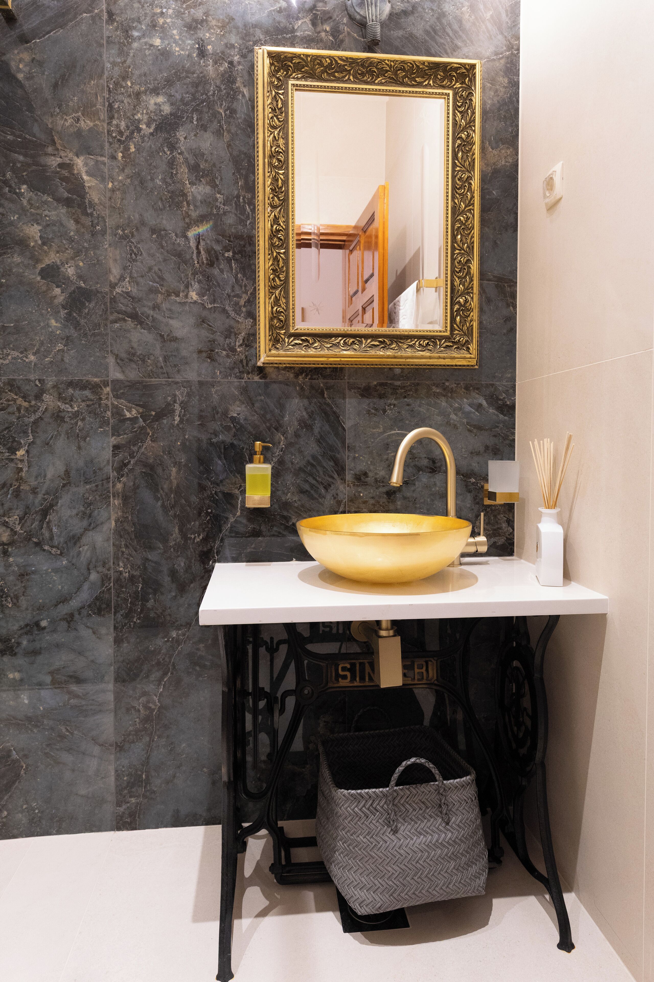Une salle de bain avec un mur en marbre, un miroir encadré d’or, un lavabo blanc avec un récipient doré, un flacon doseur noir et un vase blanc contenant des tiges de roseau.
