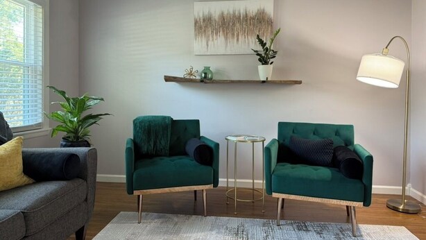A living room with two green armchairs, a grey sofa, a small round table, a floor lamp, and a painting on the wall.