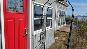 A house with a red door, a window with a metal gate, and a view of the water. - Waterfront Retreat with Loft & Harbor Views in Historic Eastport, Maine (Eastport)
