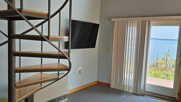 A room with a spiral staircase, a flat-screen TV mounted on the wall, a sliding glass door leading to an outdoor area, and a view of the sea. - Waterfront Retreat with Loft & Harbor Views in Historic Eastport, Maine (Eastport)