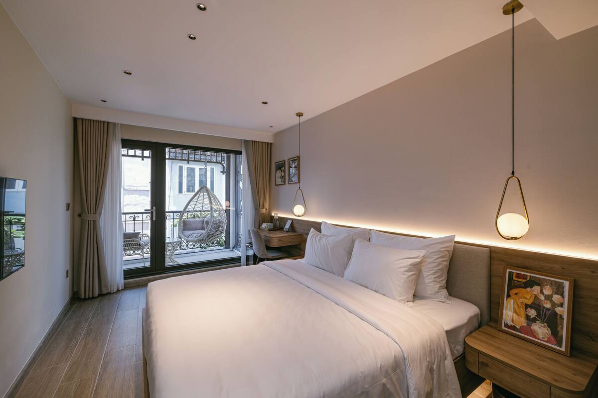 A hotel room with a large bed, a desk, a chair, and a balcony with outdoor furniture.