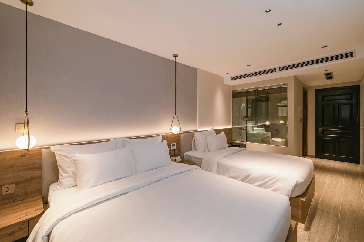 A hotel room with two beds, a wooden headboard, a nightstand, and a bathroom visible through a glass door.