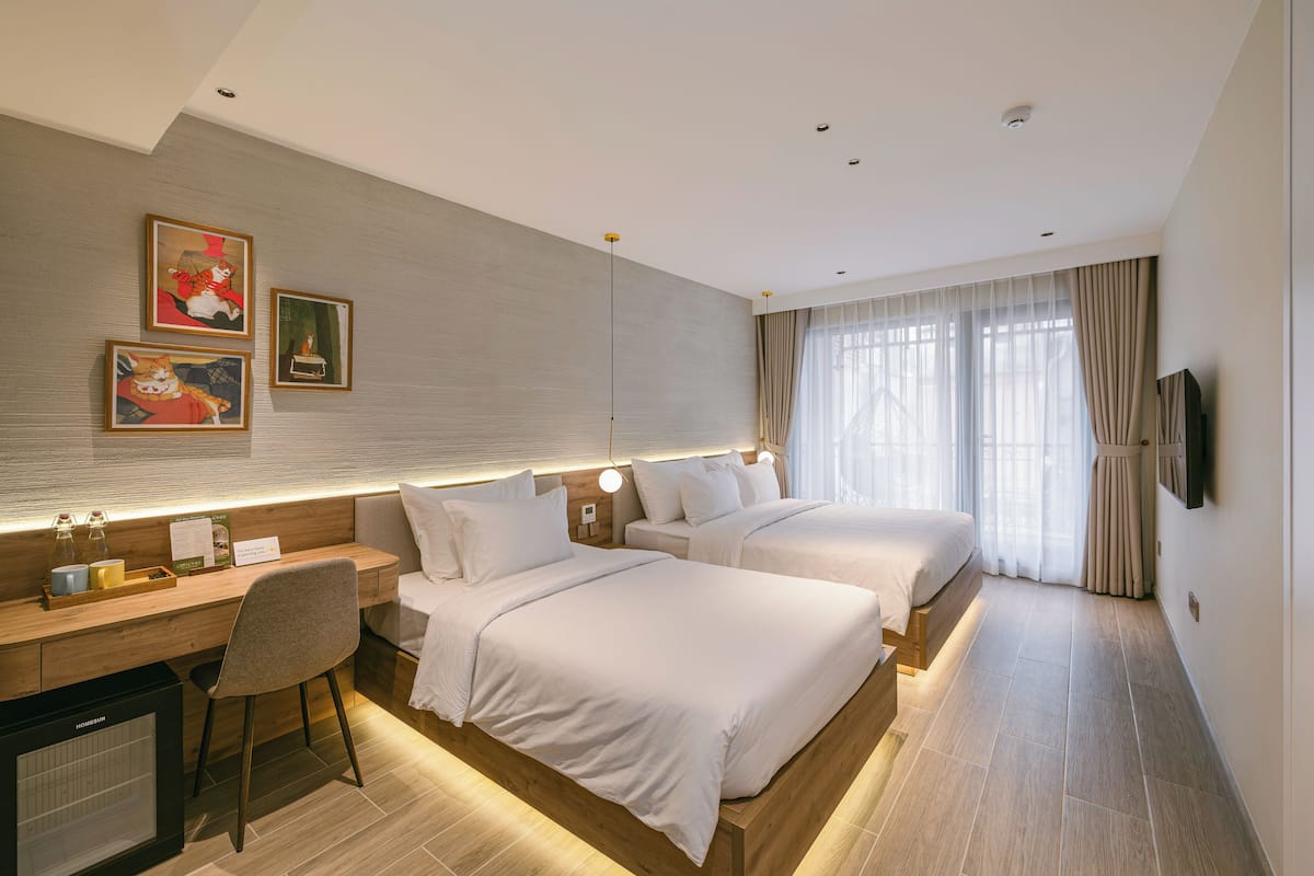 A hotel room with two beds, a desk, and a chair. There are three framed pictures on the wall, a TV mounted on the wall, and a window with curtains.