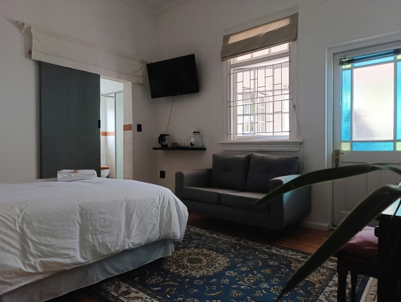 Un dormitorio con una cama, un sofá gris, un televisor en la pared y una ventana con persianas.