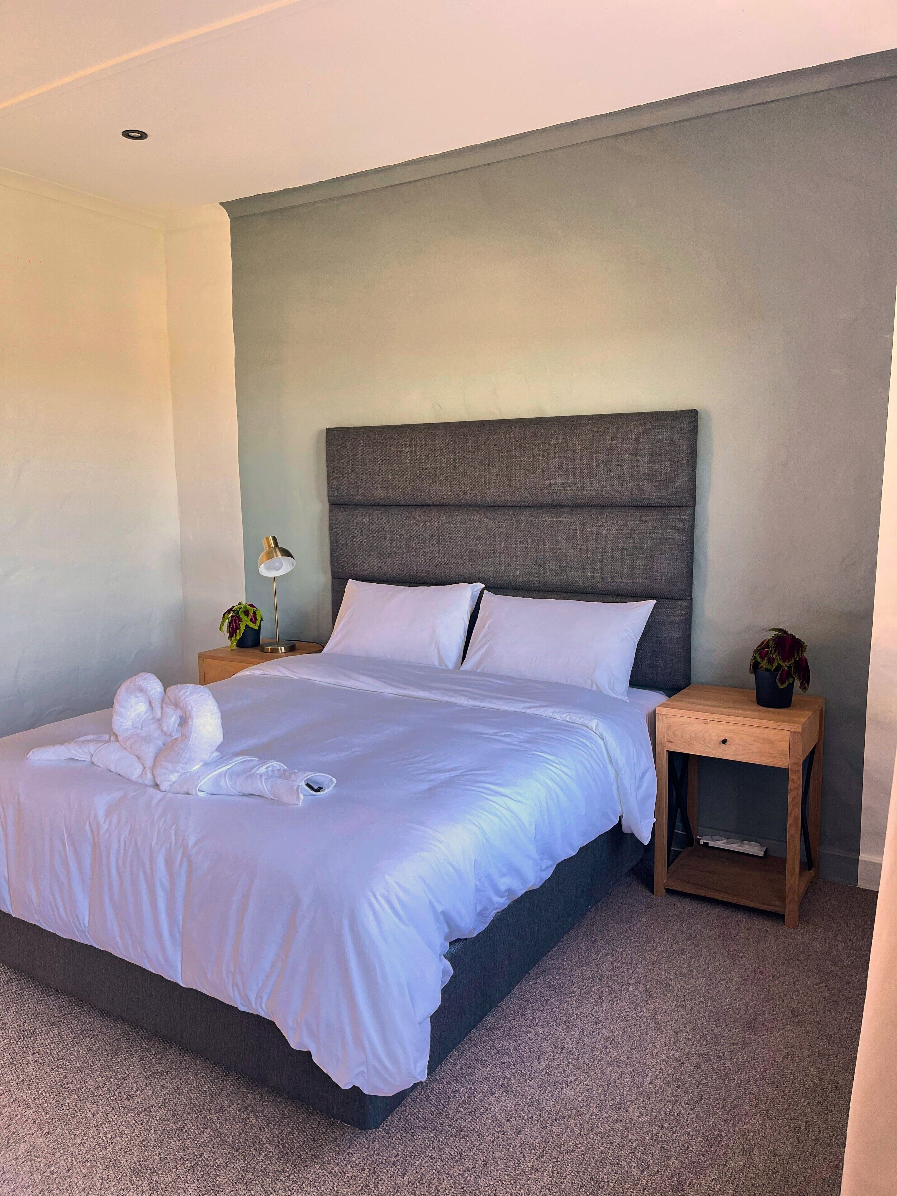 Cama bem arrumada com lençóis brancos e duas toalhas, mesa de cabeceira de madeira com luminária e planta em vaso, e cabeceira cinza.
