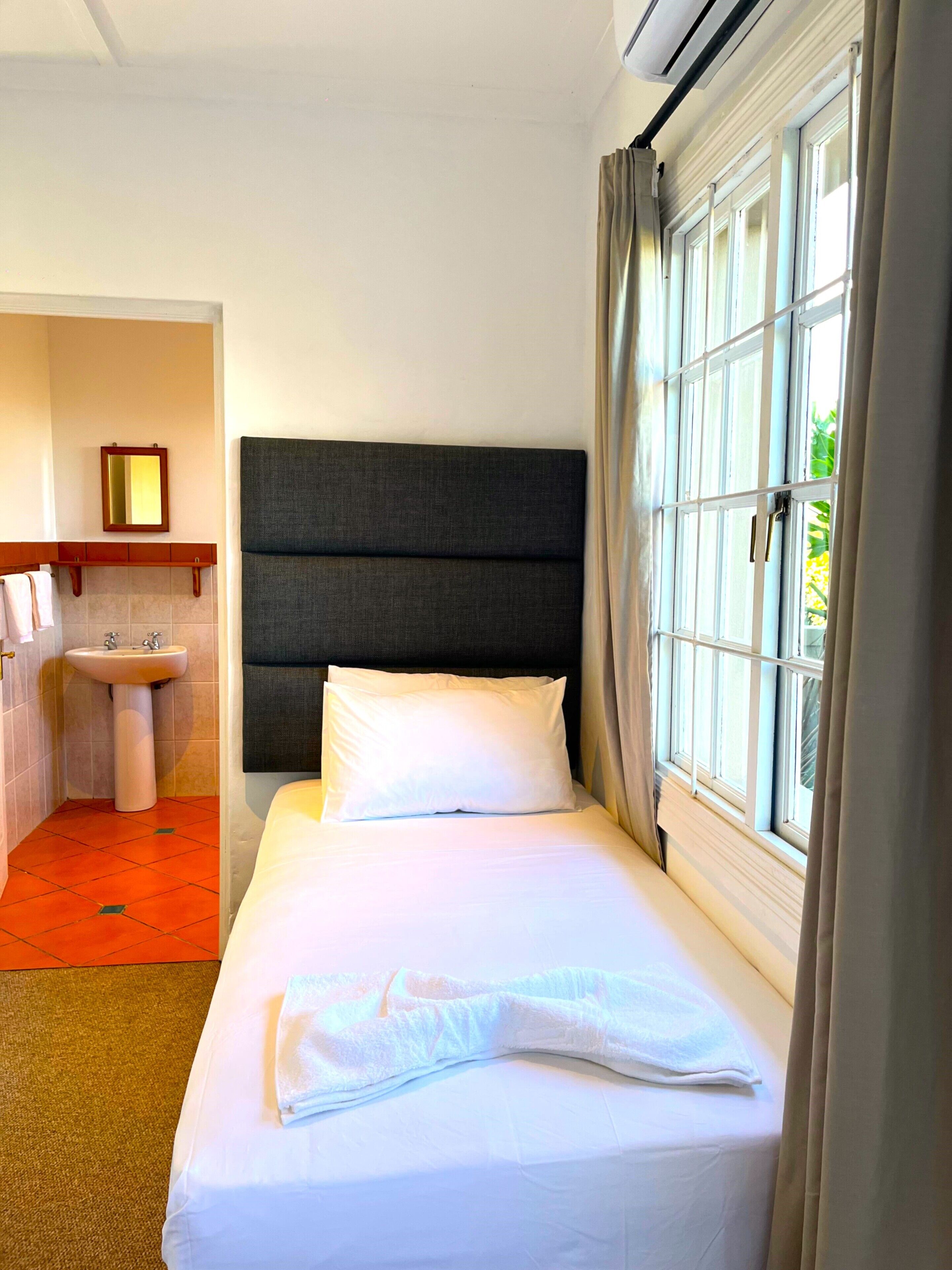A hotel room with a bed, a window with curtains, a bedside table, a mirror, and a bathroom with a sink and towel rack.